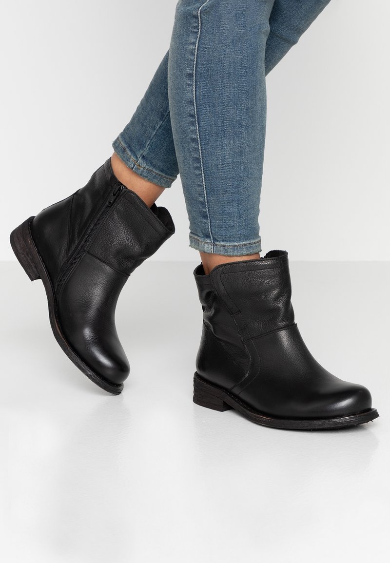 Personne portant des bottines en cuir noir à fermeture éclair latérale et talons bas empilés, associées à un jean skinny bleu retroussé.
