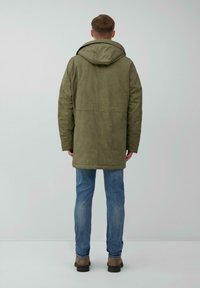 Olijfgroene parka met capuchon, met een rechte snit, gestructureerde stof en twee zijnaden, gedragen over blauwe spijkerbroek en bruine schoenen.