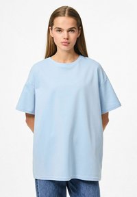 Gaiši zils pārāk liels T-krekls ar īsām piedurknēm, apaļo kakla izgriezumu un gludu kokvilnas tekstūru, apvienots ar gaišiem džinsiem.