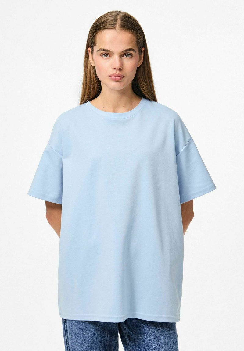 Gaiši zils pārāk liels T-krekls ar īsām piedurknēm, apaļo kakla izgriezumu un gludu kokvilnas tekstūru, apvienots ar gaišiem džinsiem.