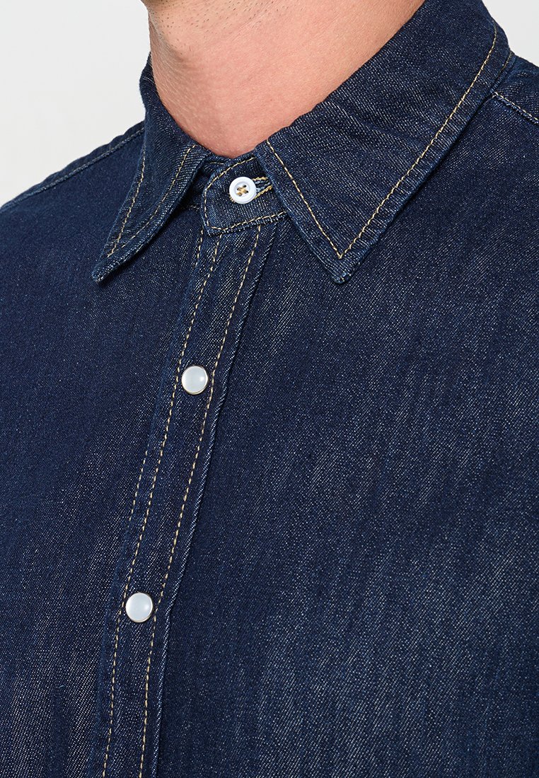Dunkelblaue Jeanshemd mit spitzem Kragen, sichtbaren Nähten und zwei Druckknöpfen am Vorderpass. Glatte Textur.