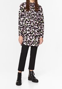 Pitkähihaisen paidan pinkki, musta ja vihreä abstrakti kuvio; edessä nappikiinnitys; käytetään mustien lyhyiden housujen ja mustien nauhallisten saappaiden kanssa.