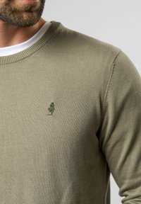 Oliwkowy sweter z prążkowanym okrągłym dekoltem. Posiada małe haftowane logo na piersi. Materiał ma gładką fakturę i lekki wygląd.
