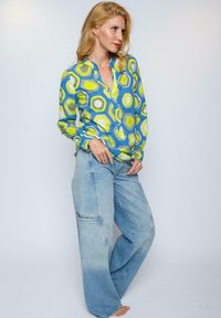 Langärmlige, blaue Bluse mit grünem und gelbem geometrischem Muster, kombiniert mit locker sitzenden, hellblauen Denim-Jeans im weiten Schnitt.