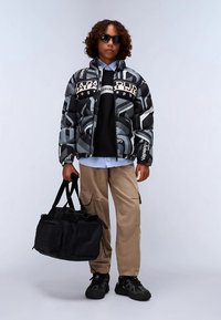 Giacca a vento nera e grigia con motivi astratti, felpa nera, camicia azzurro chiaro, pantaloni kaki, borsa nera e scarpe nere.