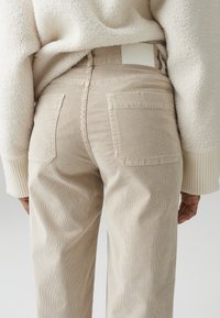 Pantaloni in corduroy beige con una superficie testurizzata, dotati di tasche posteriori e di un'etichetta bianca sulla vita, abbinati a un maglione in pile bianco.