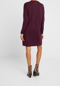 Bordeaux Strickkleid mit langen Ärmeln, rundem Ausschnitt und gerippten marineblauen Bündchen; kombiniert mit braunen Schlangenleder-Ankle-Boots mit Blockabsätzen.