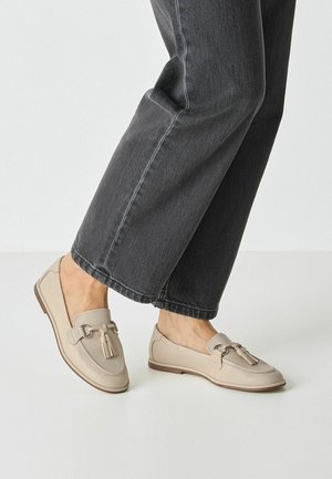 Beige Leder-Loafer mit dekorativer Quaste und Schnallendetail. Sie haben eine flache Sohle und eine abgerundete Spitze und werden zu einer dunklen Jeans getragen.