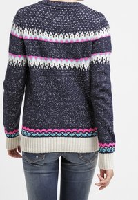 Frau trägt einen navyblauen Strickpullover mit weißen, pinken und blauen geometrischen Mustern, kombiniert mit ausgewaschenen blauen Jeans, von hinten gezeigt.