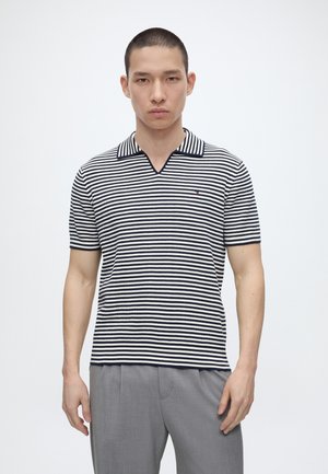 Mann trägt ein gestreiftes schwarz-weißes kurzärmeliges Polohemd und graue Hose, steht vor einem schlichten weißen Hintergrund.