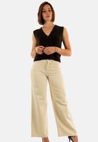 Mujer de pie vistiendo un chaleco negro sin mangas con botones, pantalones de pierna ancha beige, pulseras y tacones altos negros frente a un fondo liso.