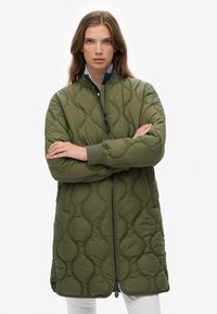 Giacca lunga in verde oliva trapuntata con colletto rialzato, zip frontale e motivo a onde testurizzato. Presenta polsini a maglia e tasche laterali.
