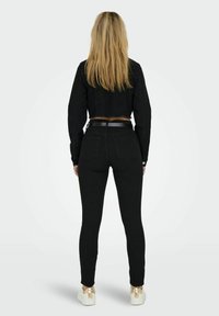 Woman with long blonde hair wearing a black cropped jacket, black high-waisted skinny jeans, black belt, and white sneakers with gold accents.