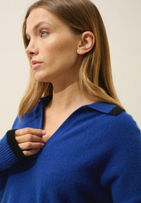 Sininen villapaita, jossa on V-pääntie, mustat yksityiskohdat kauluksessa ja hihoissa, pehmeä tekstuuri ja rento istuvuus.