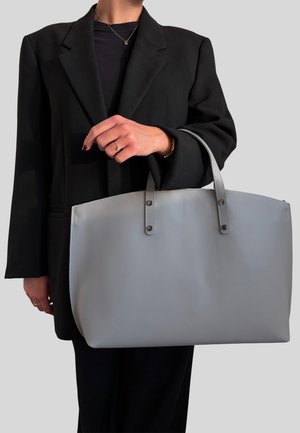 Borsa a spalla grigia chiara con due manici, caratterizzata da un design piatto e da dettagli di cucitura sottili. Indossata da una persona in un blazer nero.