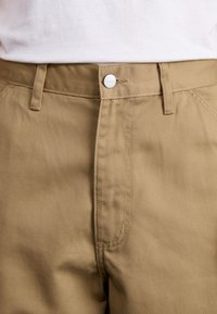 Pantaloni din bumbac bej, cu închidere frontală cu buton, curea de susținere și buzunare frontale diagonale, având o textură netedă și un design ajustat.