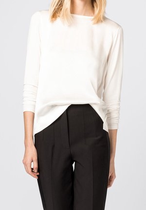 Femme portant un haut blanc à manches longues rentré dans un pantalon noir taille haute et ajusté, debout devant un fond uni.