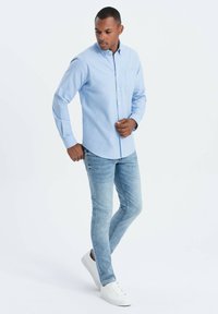 Camisa de botones azul claro con mangas largas, detalle de bolsillo, combinada con jeans azul claro y zapatillas blancas sobre un fondo liso.