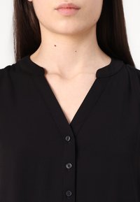 Blouse noire avec un col en V, un col court et une fermeture à boutons à l'avant avec cinq boutons noirs assortis. Texture lisse et douce.