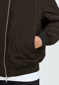 Dark brown bomber jacket with a zip front, ribbed cuffs, and a large front pocket. Smooth, slightly glossy texture. Layered over a white shirt.