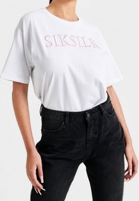 Bílá bavlněná tričko s růžovým vyšitým nápisem "SIKSILK", spárované s černými džínami s vysokým pasem, které nabízejí volný střih a jednobarevný vzhled.