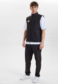 Schwarze ärmellose Weste mit hohem Kragen, ausgestattet mit zwei Reißverschlusstaschen und einem Logopatch, über einem weißen Kurzarmhemd und schwarzen Hosen getragen.