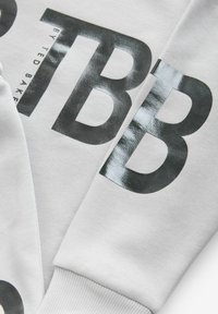 Sudadera gris con letras impresas en negro en negrita "TB" y "B" con un acabado brillante, puños acanalados y textura suave.