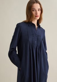 Abito camicia blu navy in tessuto testurizzato con colletto a bottoni, davanti plissettato e maniche lunghe con polsini. Presenta tasche laterali.