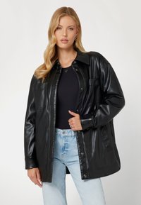 Giacca nera in similpelle con vestibilità morbida, chiusura a bottoni e due tasche frontali, abbinata a un top nero e jeans azzurri chiari.