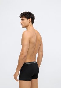 Homem com cabelo encaracolado e escuro, sem camisa, vestindo boxers pretos da Calvin Klein, de perfil contra um fundo claro e simples.