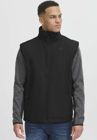 Črna softshell brezrokavnik z sivimi rokavi, ki spominjajo na pulover, visokim ovratnikom, zapiranjem na zadrgo in dvema stranskima žepoma. Ima logo na prsih.