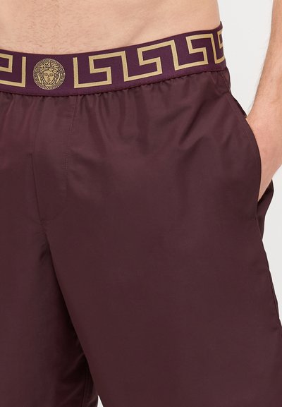 Σκούρο κόκκινο μαγιό με ελαστική ζώνη διακοσμημένη με χρυσό μοτίβο που απεικονίζει ένα μετάλλιο. Πλευρές τσέπες και απαλό, ελαφρύ υλικό.