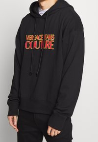 Svart hoodie med framficka, dragsnöre och ribbade ärmslut. Har broderad text i rött och gult "VERSACE FANS COUTURE" på bröstet.