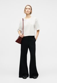 Frau trägt ein weißes, weit geschnittenes Kurzarmoberteil, schwarze Hosen mit weitem Bein, schwarze Schuhe und trägt eine dunkelbraune Umhängetasche vor weißem Hintergrund.