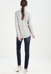 Grauer Pullover mit hohem Kragen, gerippten Bündchen und einem etwas längeren Rücken. Kombiniert mit dunklen Jeans und silbernen Sneakers. Glatte Textur.