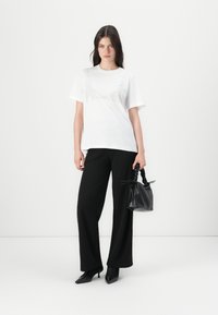 White t-shirt with a textured pattern on the front, paired with black wide-leg pants and a small black handbag. Model stands confidently.
