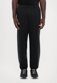 Calças de atleta pretas com uma cintura elástica, bolsos laterais e um corte descontraído. Combinadas com ténis pretos com detalhes em texto branco.