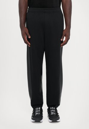 Black athletic pants with an elastic waistband, side pockets, and a relaxed fit. Paired with black sneakers featuring white text accents.