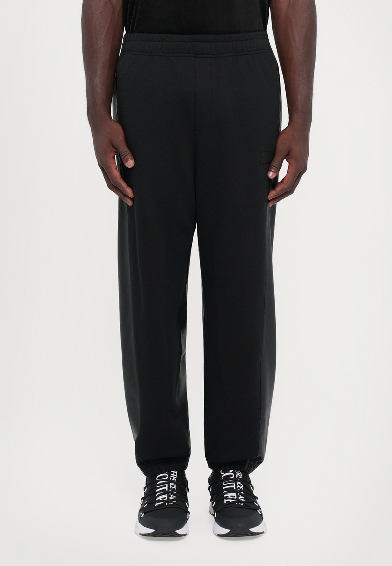 Calças de atleta pretas com uma cintura elástica, bolsos laterais e um corte descontraído. Combinadas com ténis pretos com detalhes em texto branco.