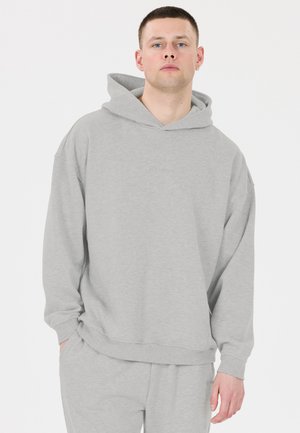 Young man with short hair wearing a light gray hoodie and matching sweatpants, standing with one hand in pocket on white background.