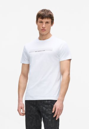 Ung mand med kort hår iført en hvid Karl Lagerfeld Jeans T-shirt og sorte mønstrede bukser, stående foran en ensartet baggrund.