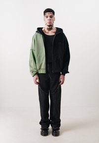 Oversized hoodie in green and black cotton, paired with black pants featuring frayed edges. Model stands on a plain white background.