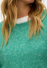 Cheveux blonds tombant sur un pull vert en tricot superposé à un t-shirt blanc à col rond.