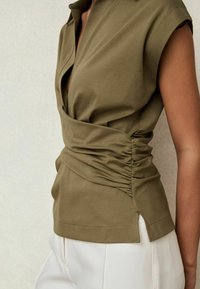 Top sin mangas de color verde oliva con detalle fruncido en el lateral, combinado con pantalones blancos de cintura alta, mostrando parcialmente la parte superior del torso y el brazo.