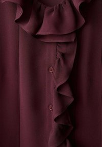Blouse bordeaux avec un col volanté, des fermetures à boutons et un tissu lisse et texturé. Les volants se prolongent le long de la patte d'imposte.