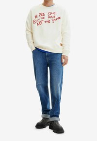 Sweatshirt crema con texto bordado en rojo, camisa azul debajo, combinado con jeans azul claros y zapatos gruesos negros. Un atuendo sencillo y casual.