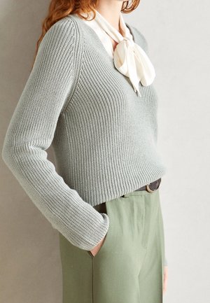 Femme portant un pull en maille gris clair, une écharpe crème nouée en nœud papillon et un pantalon vert pâle taille haute avec une ceinture noire, main dans la poche.