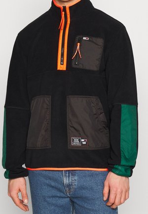 Schwarzer Fleece-Pullover mit orangefarbener Reißverschlussblende, grünen Ärmelfeldern, mehreren Reißverschlusstaschen und Frontlabel, getragen zu blauen Jeans.