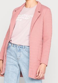 Blazer rosa chiaro con texture, colletto a scollatura, bottone singolo e tasche laterali, indossato sopra una maglietta grafica rosa chiaro e jeans a vita alta.