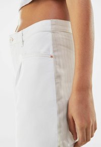 Pantalones de mezclilla blancos con una textura suave, bolsillo lateral con un acento de herrajes en oro rosa y un panel a rayas beige en uno de los laterales.